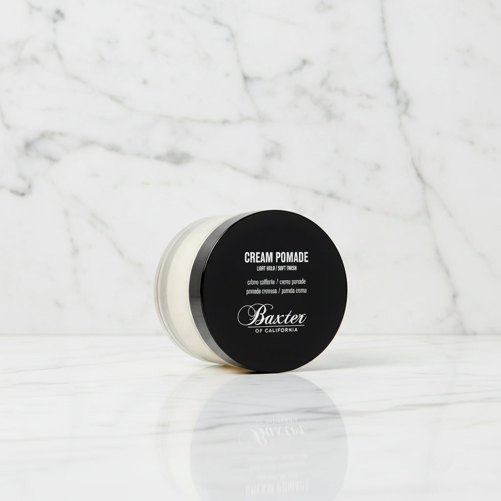 A jar of Baxter of California Cream Pomade, Light Mud / Soft Finish, featuring a black lid with white text and a clear container showing the off-white cream product inside, set against a plain white background.