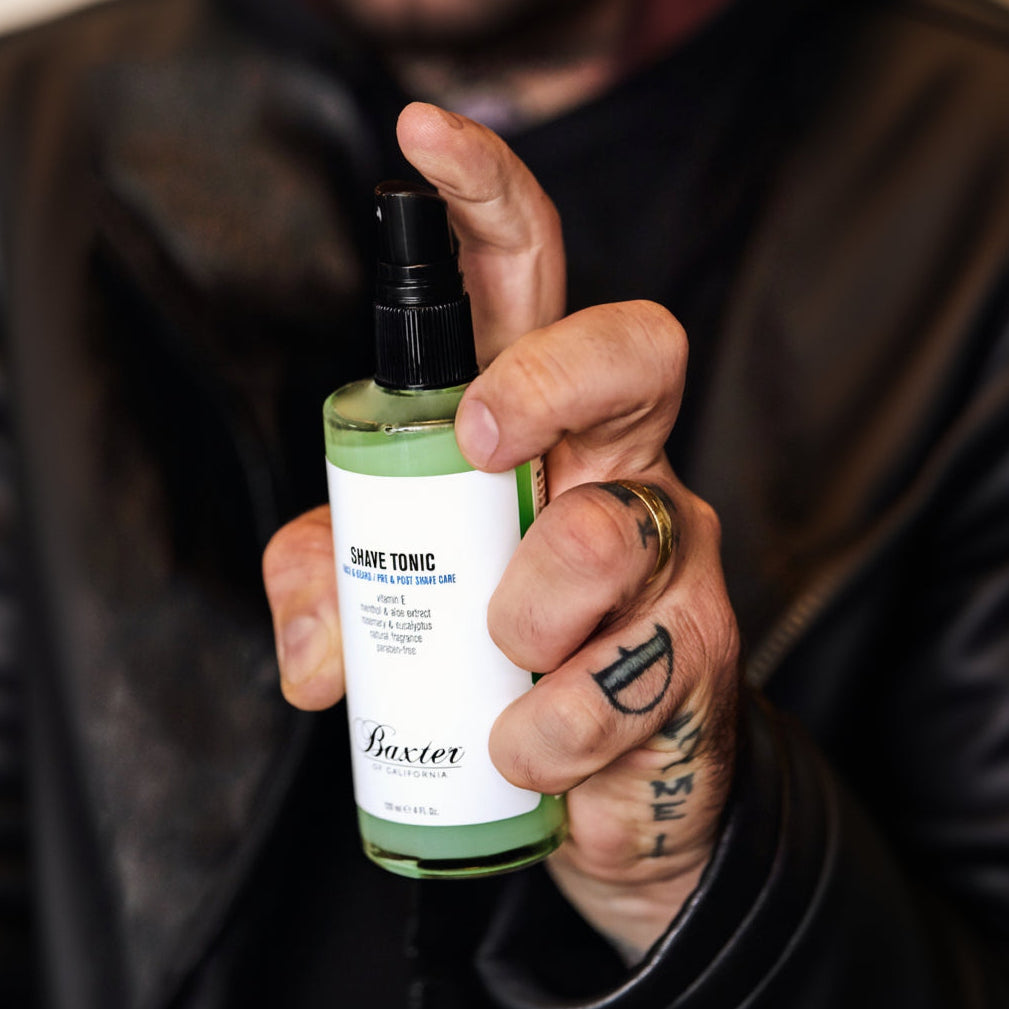 A hand holding a bottle of Baxter Men Shave Tonic with a black spray top and a white label, set against a dark, blurred background.