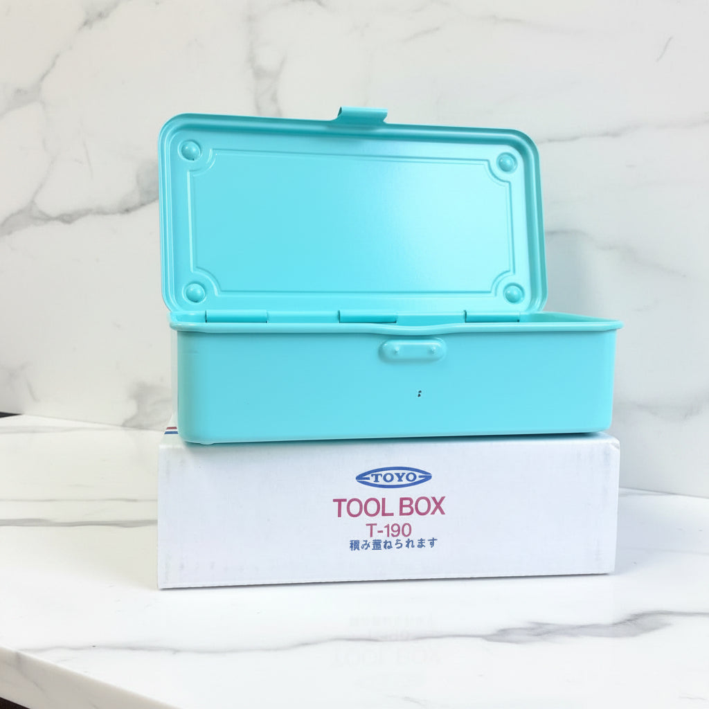 A light blue plastic tool box with its lid open, stacked on top of its white branded cardboard box. More identical boxes are stacked in the background on a white counter.